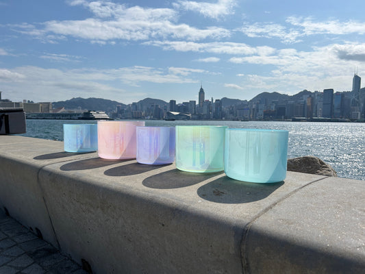 Crystal singing bowls in five different colours, placing near the sea shore. Blue sky and shimmering ocean.