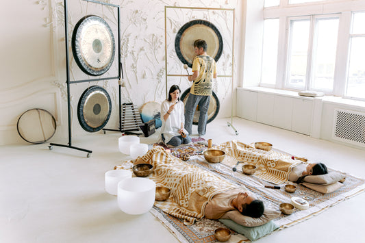 This is a sound bath healing section. Two receiver lying down, surrounding by different kinds of singing bowls and gongs. A men is playing the gongs and a women is using a palo santo to clean the space.